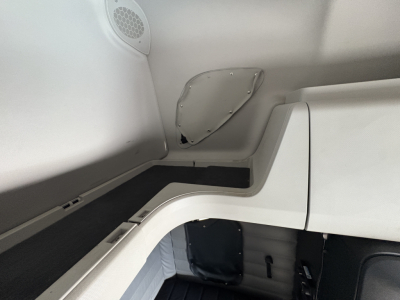 Truck cab interior corner with light gray panels and a rounded access panel; speaker grill visible on upper left; curved doorway and floor edge.