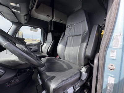 Truck cab interior showing driver seat, steering wheel, dash, and overhead storage in a 2023 Freightliner Cascadia 126 (view from doorway).