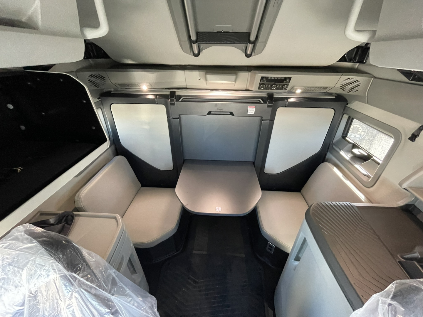 Cab interior of a new Volvo VNL64T860 sleeper, front seating area with a tabletop, light gray upholstery, overhead storage, and multipanel windows.