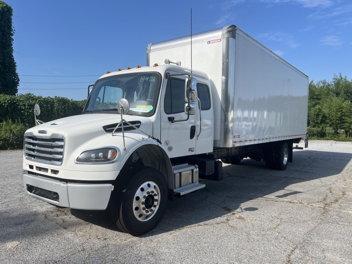 Freightliner M2 106 Plus box truck in service bay with attached box trailer; cab shows side mirror, front grille, and wheel; white exterior; boxed cargo area.