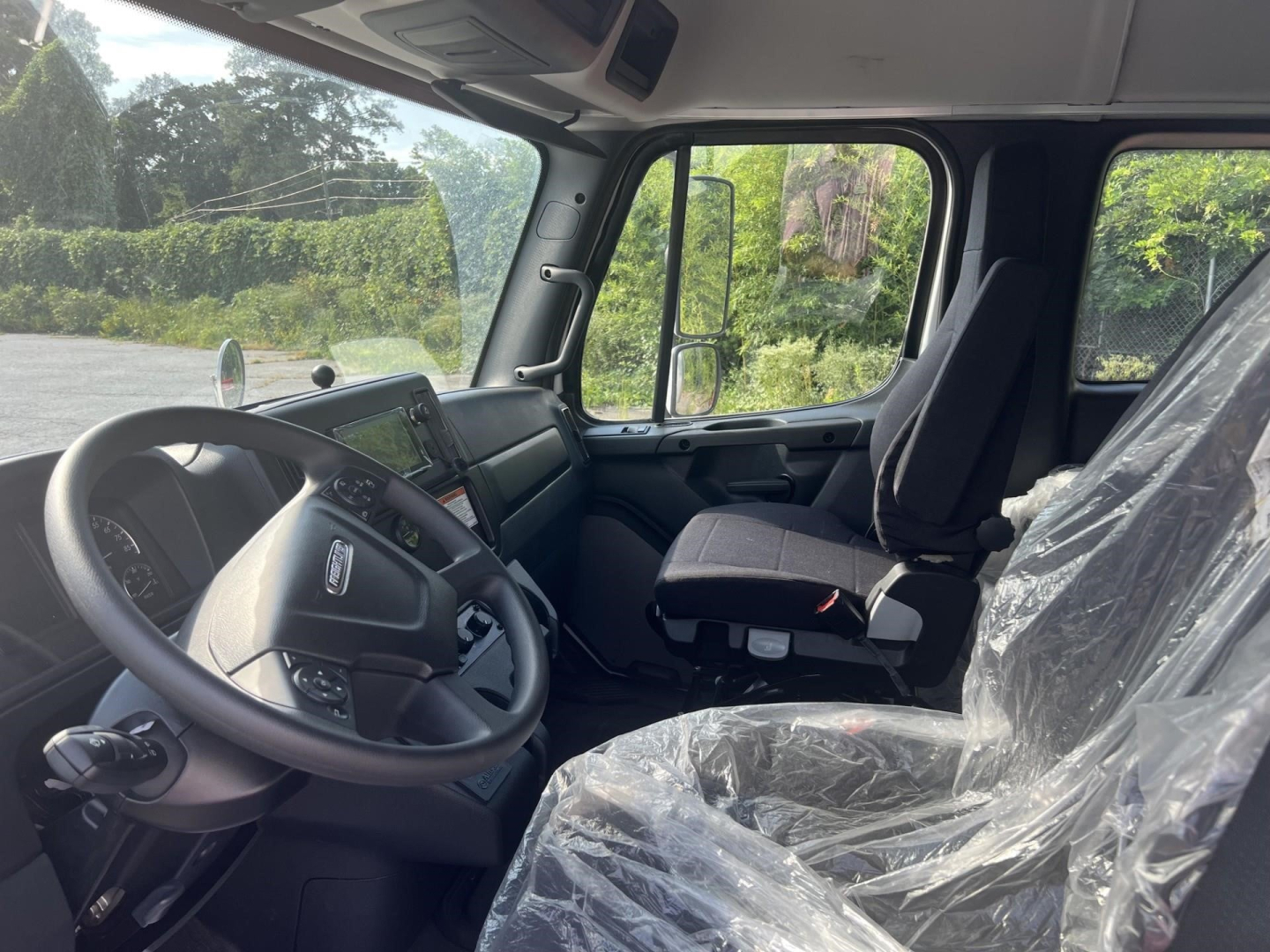Cab interior of a 2027 Freightliner M2 106 Plus box truck showing steering wheel, dashboard, driver seat covered in plastic, and passenger seat with protective wrap.