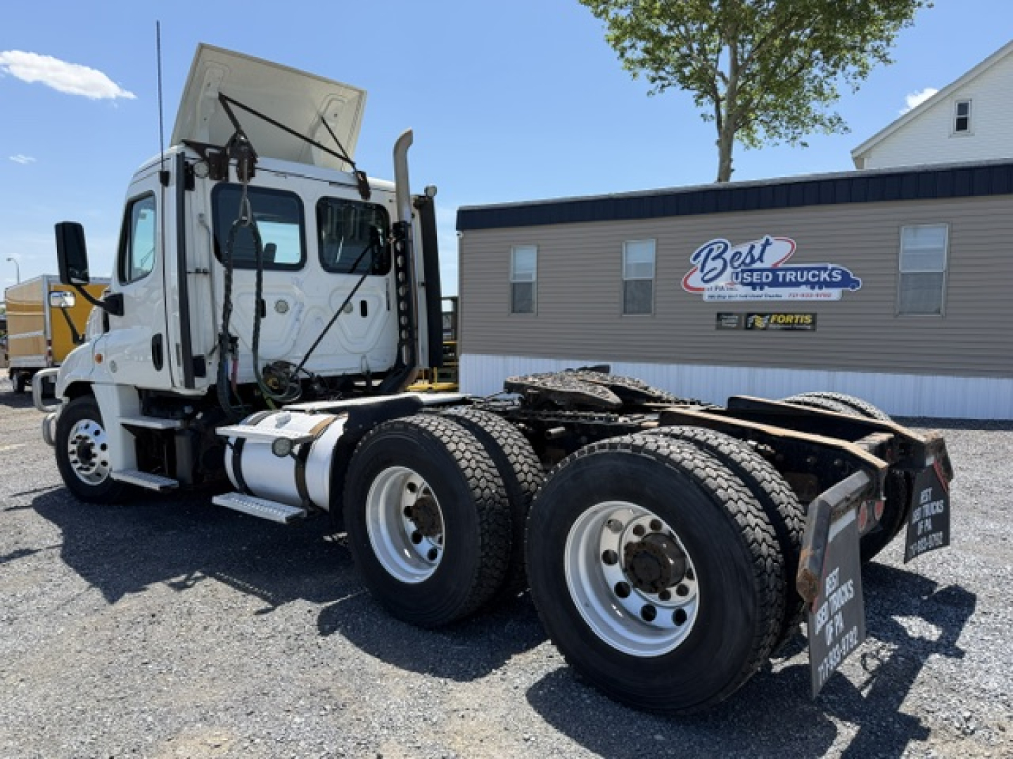 Truck chassis with cab of a Freightliner Cascadia CA125DC shown from side-rear angle, empty fifth wheel, dual rear axles, white cab, Best Used Trucks sign in background.