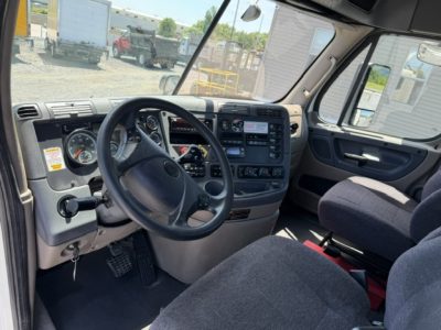 Cab interior of a Freightliner Cascadia CA125DC showing steering wheel, dashboard with gauges, center console, and two seats.