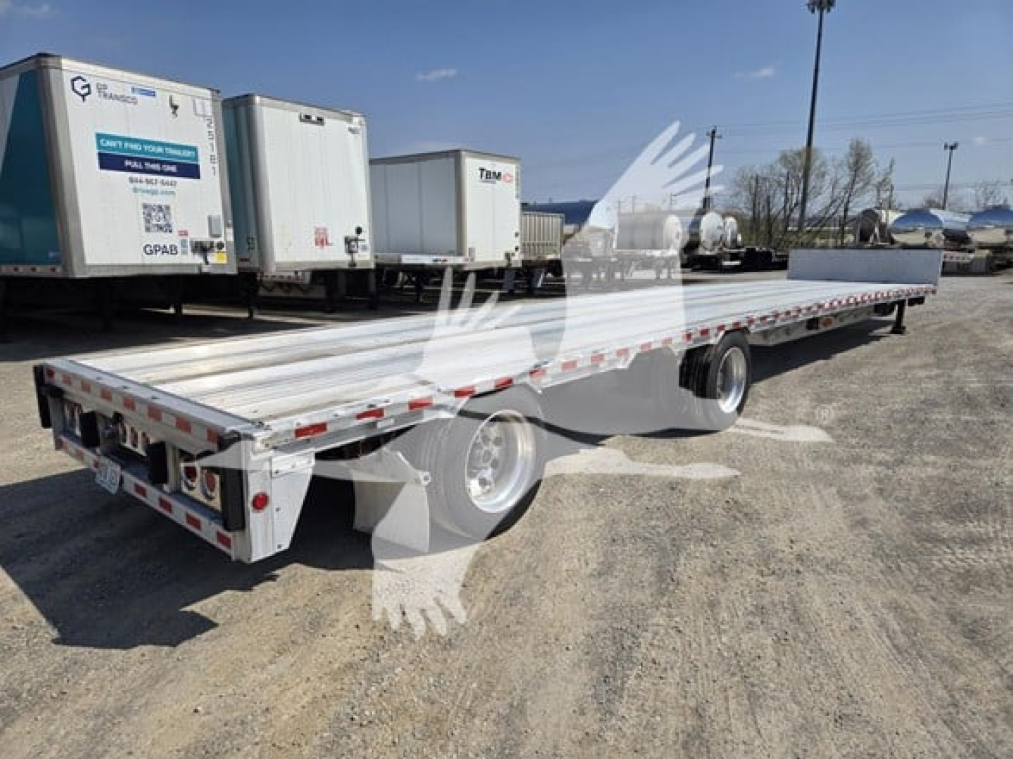 Flatbed trailer with drop deck showing long aluminum bed and red/white safety reflectors; rear lights and dual axles visible; background includes trailers and yard equipment.