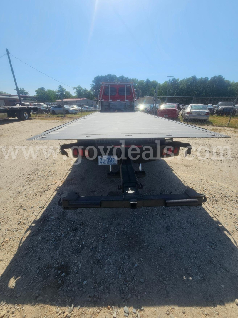 Flatbed tow truck rear with flatbed extended, VIN plate visible, no license plate, driveway yard with other vehicles.