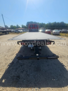 Flatbed tow truck rear with flatbed extended, VIN plate visible, no license plate, driveway yard with other vehicles.