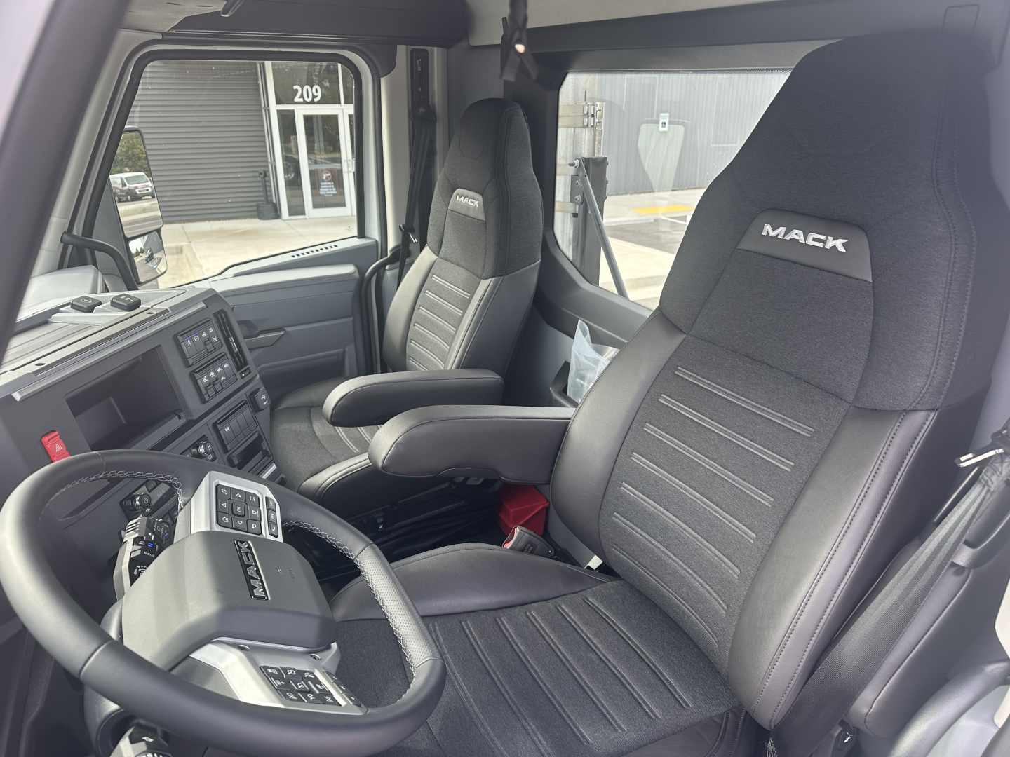 Cab interior of a new Mack PR64T daycab showing black fabric seats with Mack logo, steering wheel, and dashboard controls.