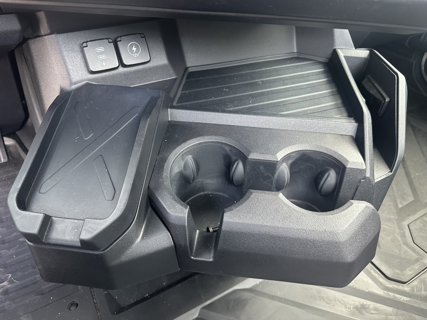 Truck interior center console with cup holders and storage compartments; visible are two buttons near the dash and a textured floor mat.