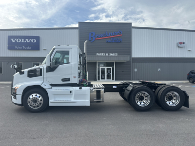  chassis of a white Mack PR64T truck showing cab with spare wheel, dual rear axles, and a business storefront in background with signs; visible text includes Volvo Volvo Trucks and Bracken Truck & Equipment Parts & Sales signage.