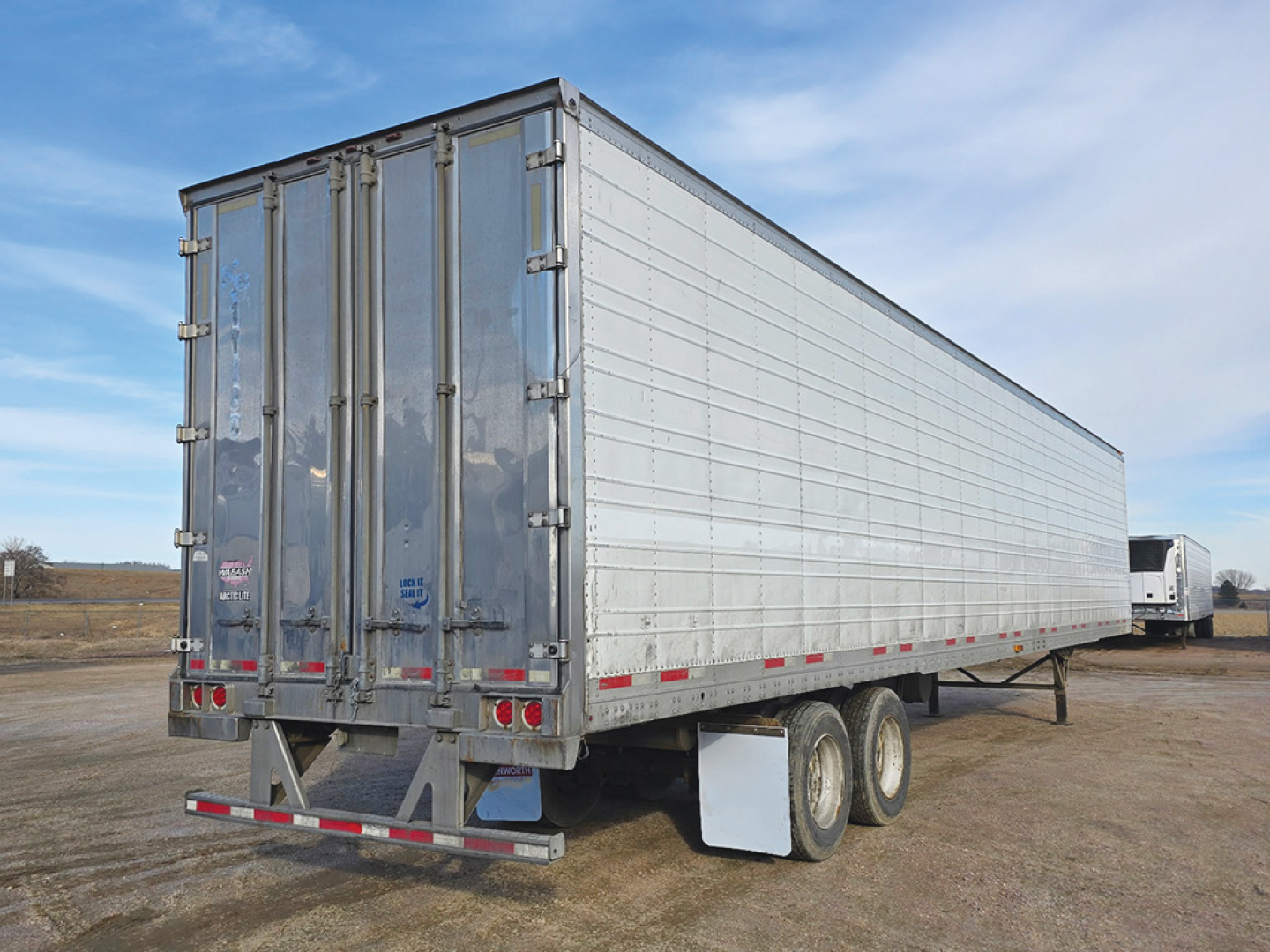 Trailer rear and side view showing a long aluminum reefer trailer with dual rear doors and twin axle setup. Visible text on the rear door hardware and lights.
