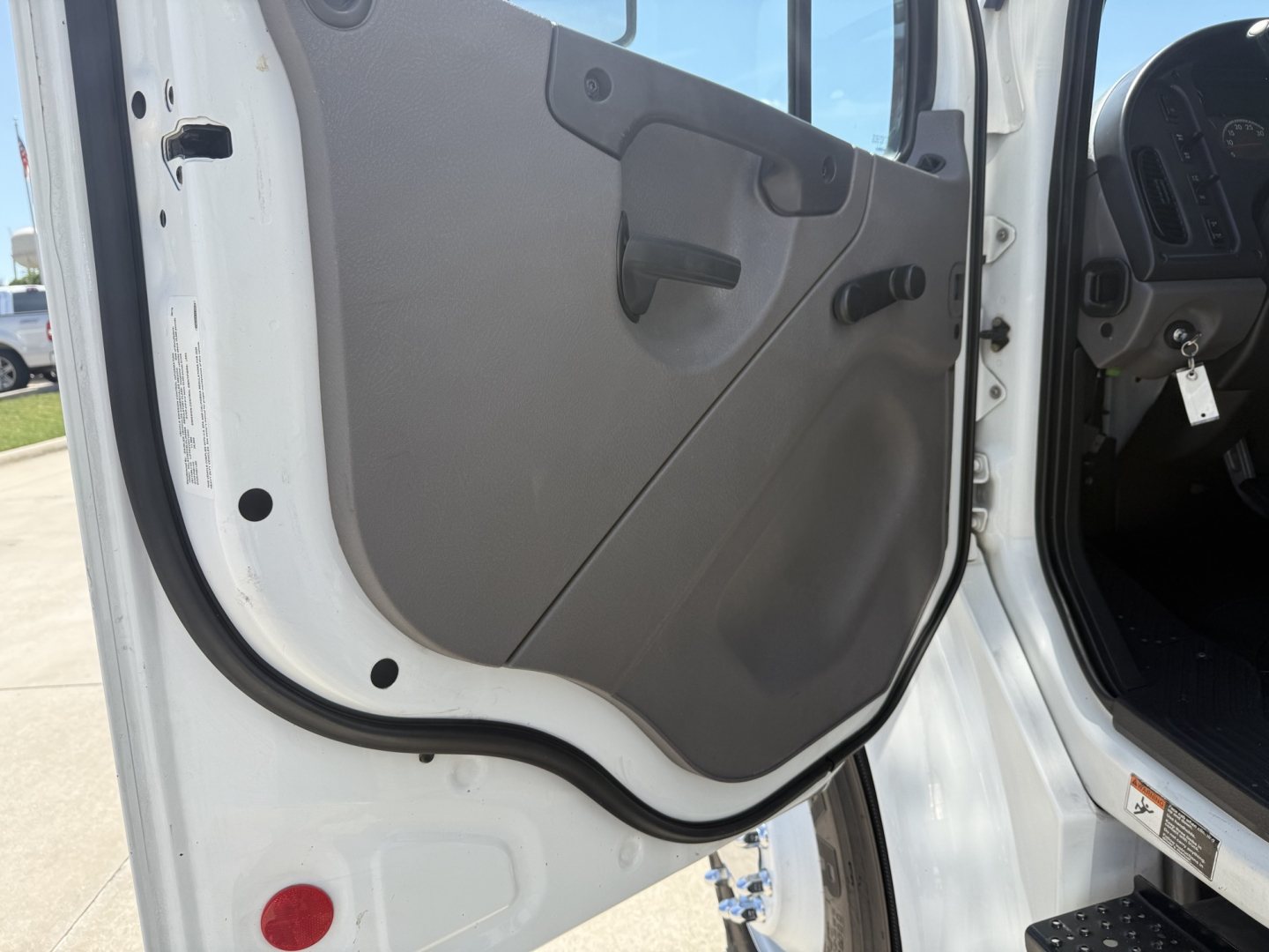 Truck cab door interior panel with manual window crank and handle; part of Freightliner M2 cab visible; light gray door panel, black window crank, beige/gray interior trim.