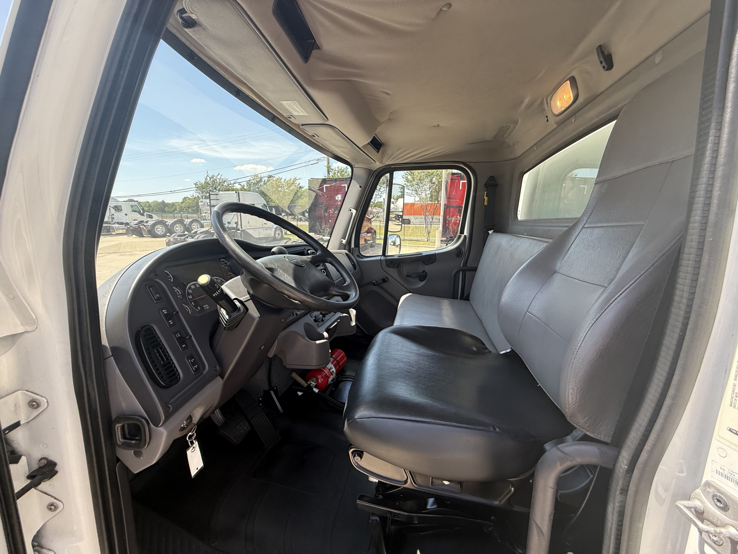 Truck cab interior showing steering wheel, driver seat, passenger seat, and dashboard of a Freightliner M2 with basic gauges and controls visible.