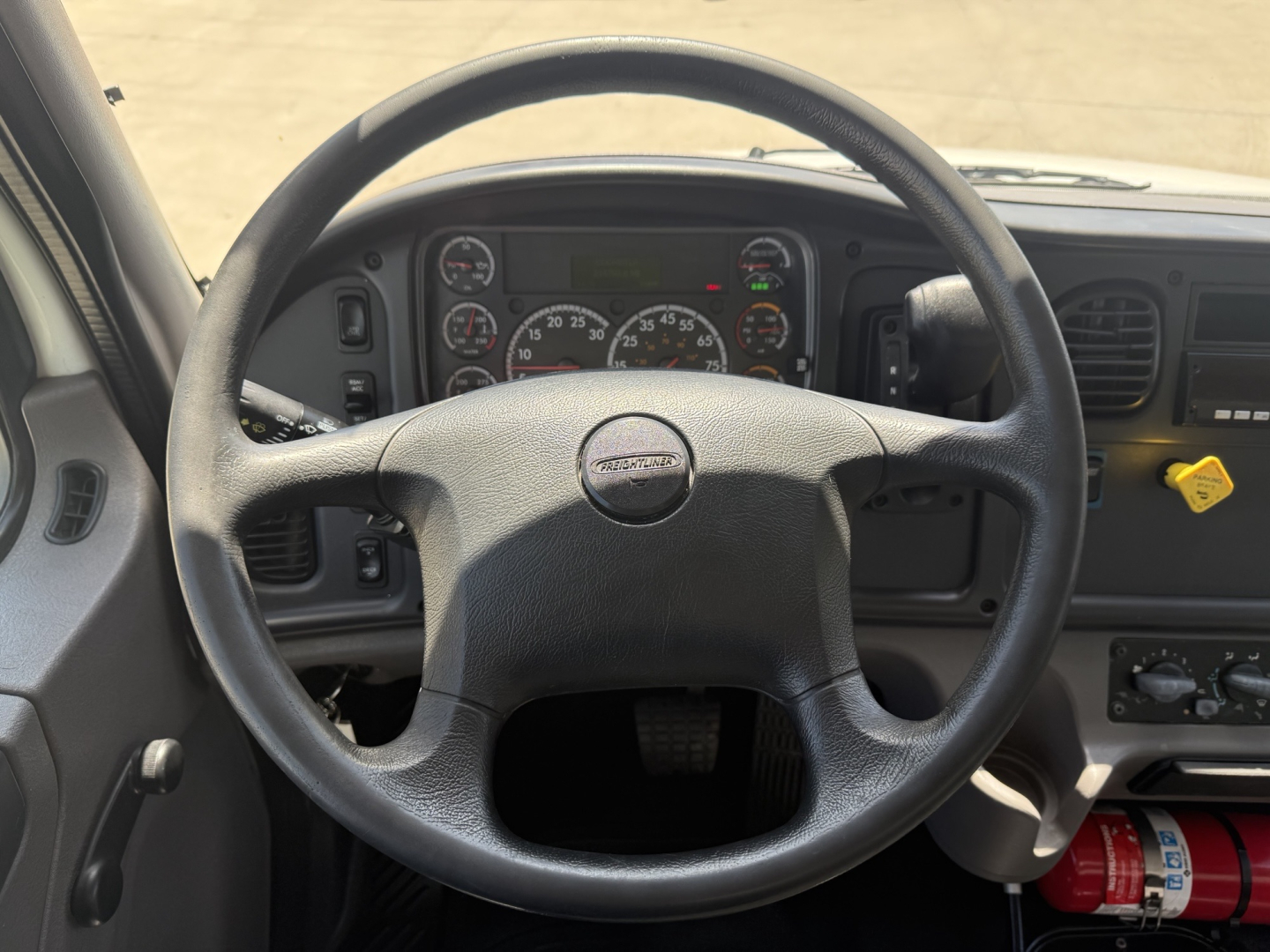 Truck cab interior showing steering wheel and instrument cluster from a Freightliner M2; dashboard gauges visible with a yellow ignition tag at right; general view of steering area.