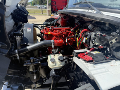 Freightliner M2 engine bay showing red engine components, black hoses, battery, and various wiring harnesses.