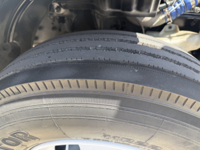 Truck wheel and tire close-up with suspension and blue hoses in background; no legible labels or text visible on tire face.