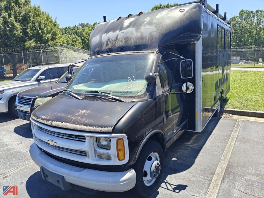 Box truck with a raised cargo area on a Chevrolet Express G3500 chassis, black cab and box, front grille and headlamps visible, parking lot background.