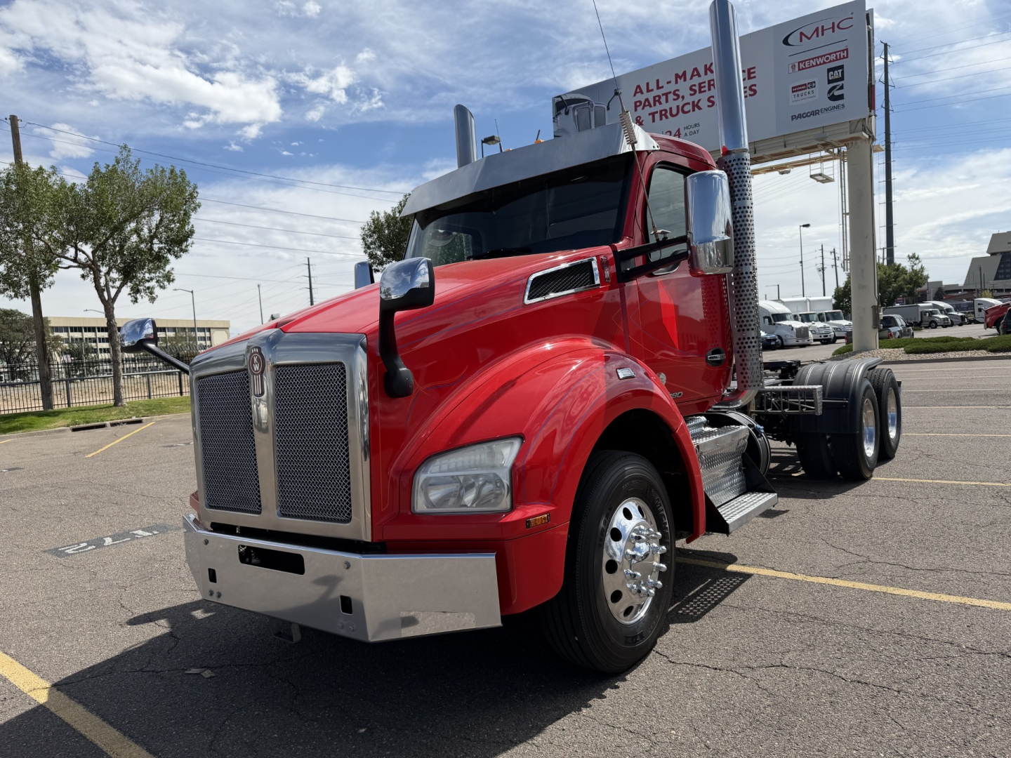 Kenworth T880 conventional daycab truck in red, front-left view, chrome grille, large silver bumper, dual rear wheels, dealership lot in background.