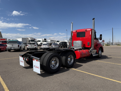 Kenworth T880 tractor in a parking lot, red cab, dual rear axles, empty flatbed area, with chrome exhaust stacks and registration signs on mudflaps.
