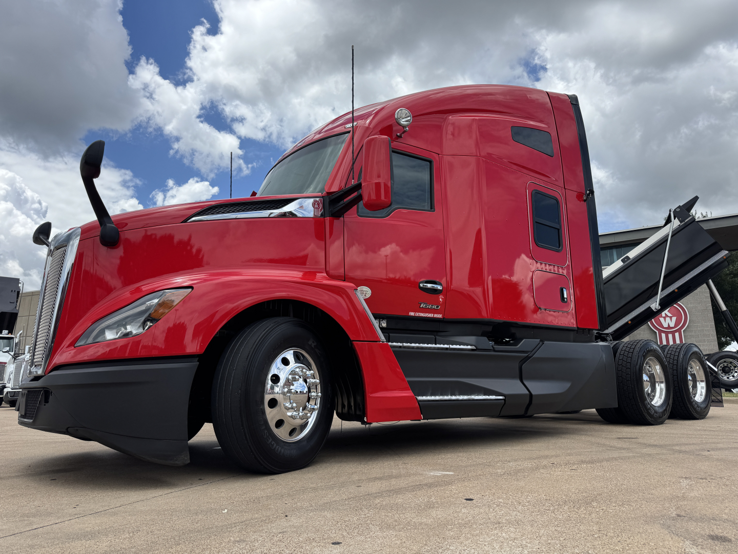 2013 Kenworth T680 red sleeper tractor front left view showing side mirror, window, wheel, and dual rear axles with a dump bed spray. Image contains no legible branding text visible.