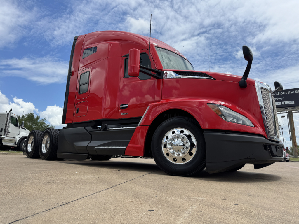Kenworth T680 conventional sleeper truck side view in red with large chrome wheel hubs and black lower body, under blue sky.