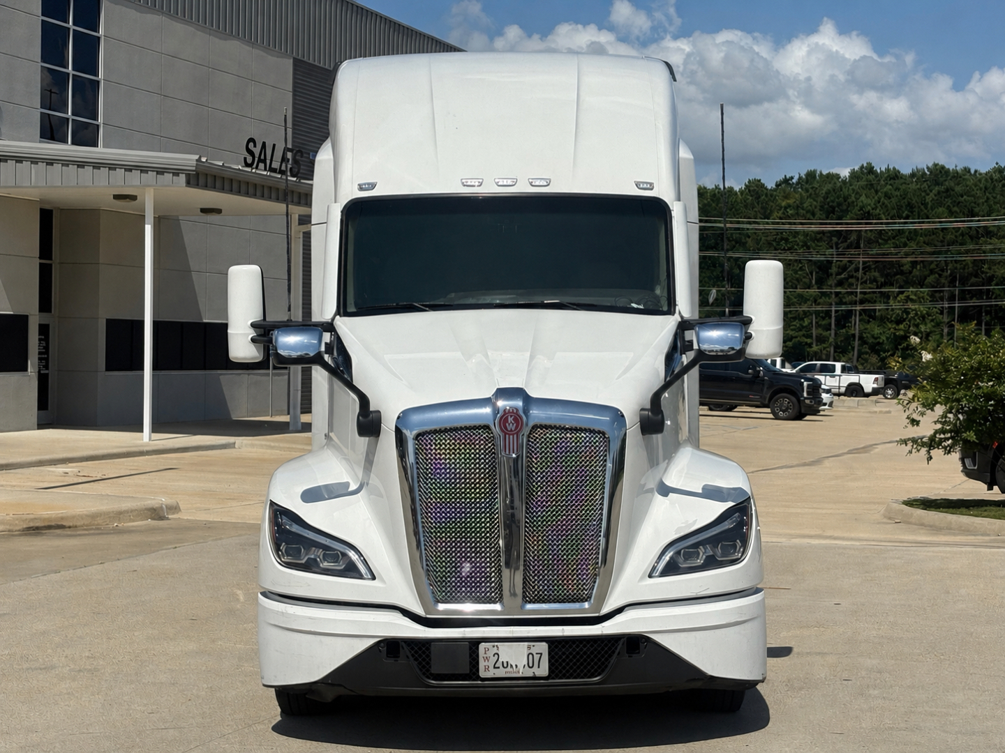 Kenworth T680 front view showing large grille with chrome surround, dual wiper-less windshield, rectangular headlights, white cab, side mirrors, and bumper with license plate area.