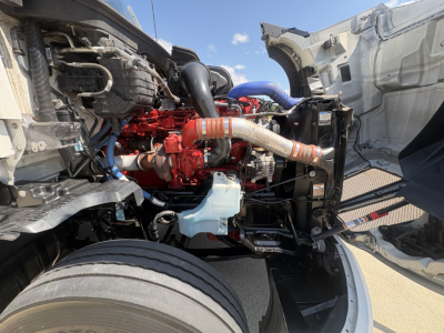 Kenworth T680 engine bay view showing red diesel engine, intake piping, blue coolant reservoir, and surrounding components; cab open for inspection.