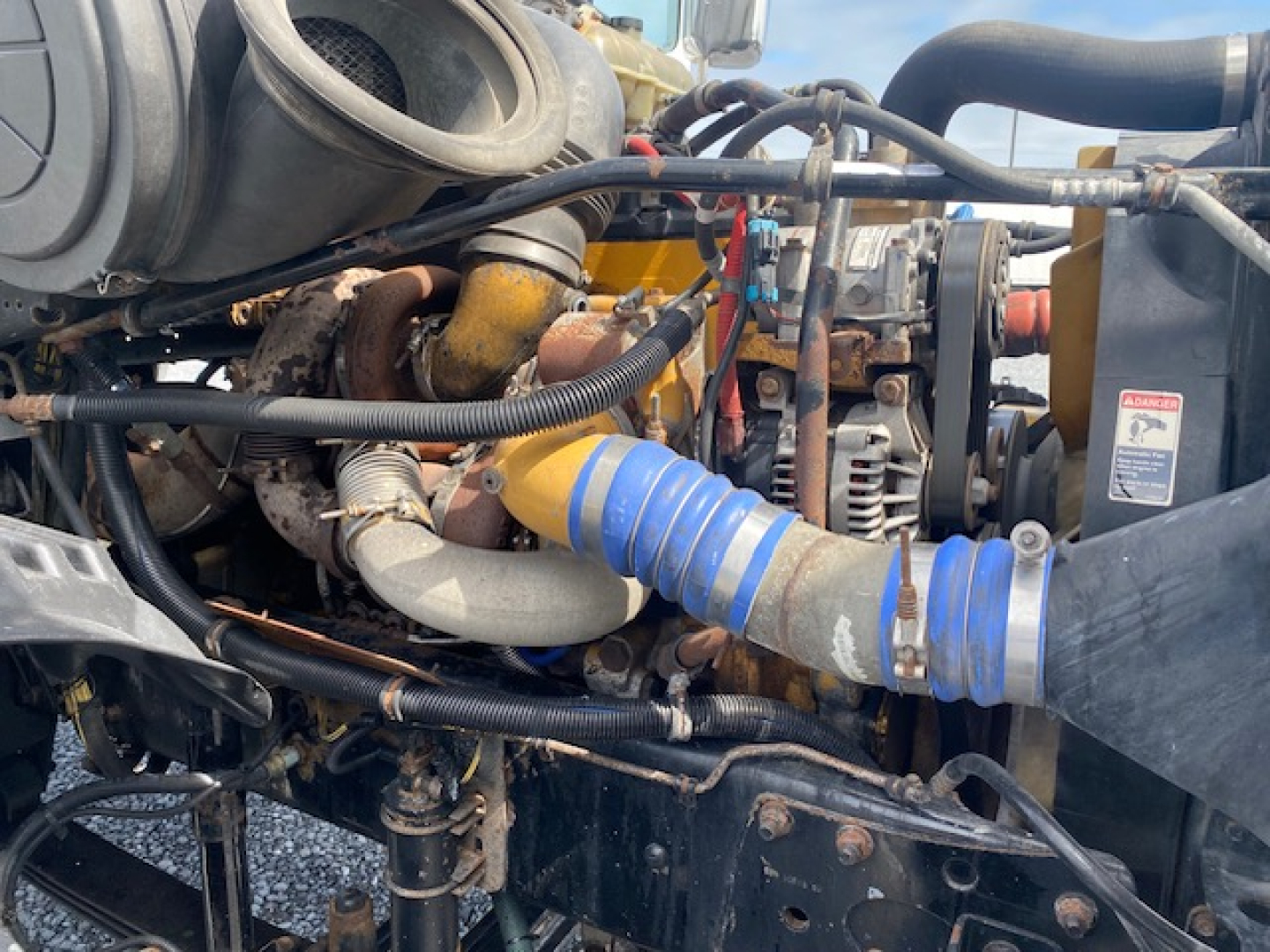 Industrial diesel engine bay with blue corrugated hose, various metal pipes, wiring, and a turbocharger visible. Labels and components partially obscured.