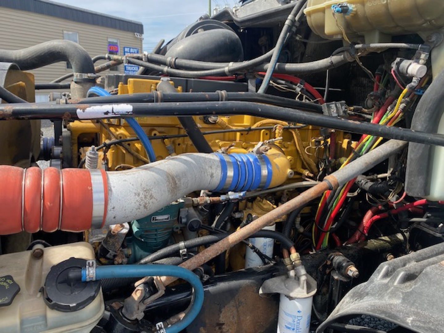 Diesel truck engine bay showing yellow engine, blue hoses, orange coolant hose, and numerous cables in a Freightliner Columbia CL120ST.