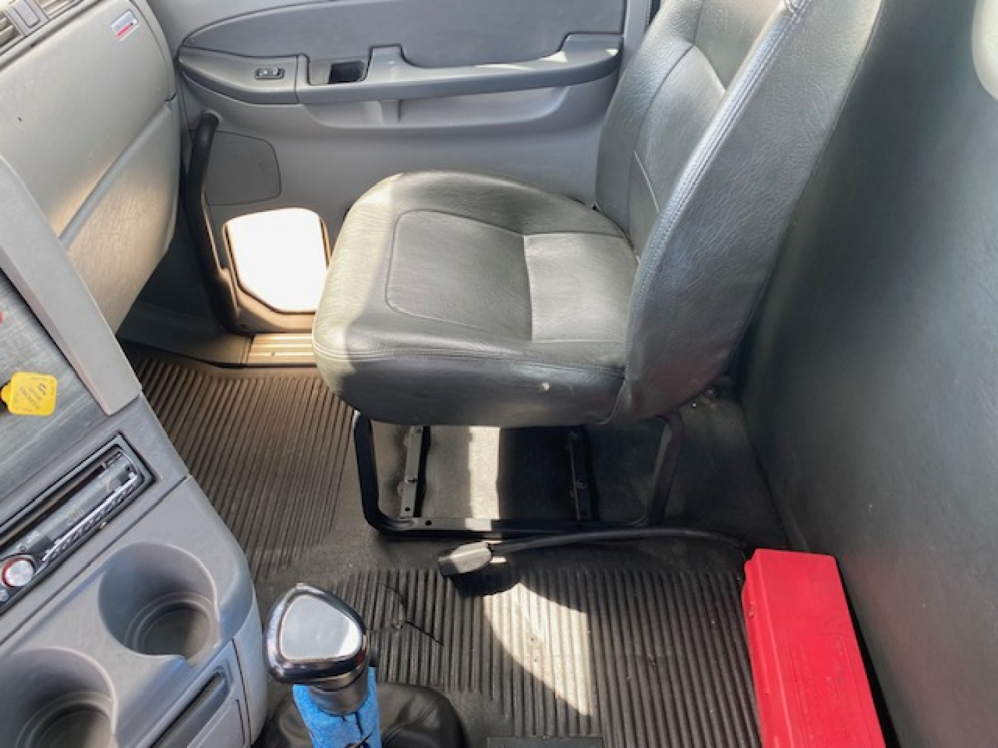 Cab interior of a Freightliner truck showing driver seat, center console, and door with control panel. Light entering from window, rubber floor mats, red toolbox on floor behind seat.