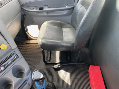 Cab interior of a Freightliner truck showing driver seat, center console, and door with control panel. Light entering from window, rubber floor mats, red toolbox on floor behind seat.