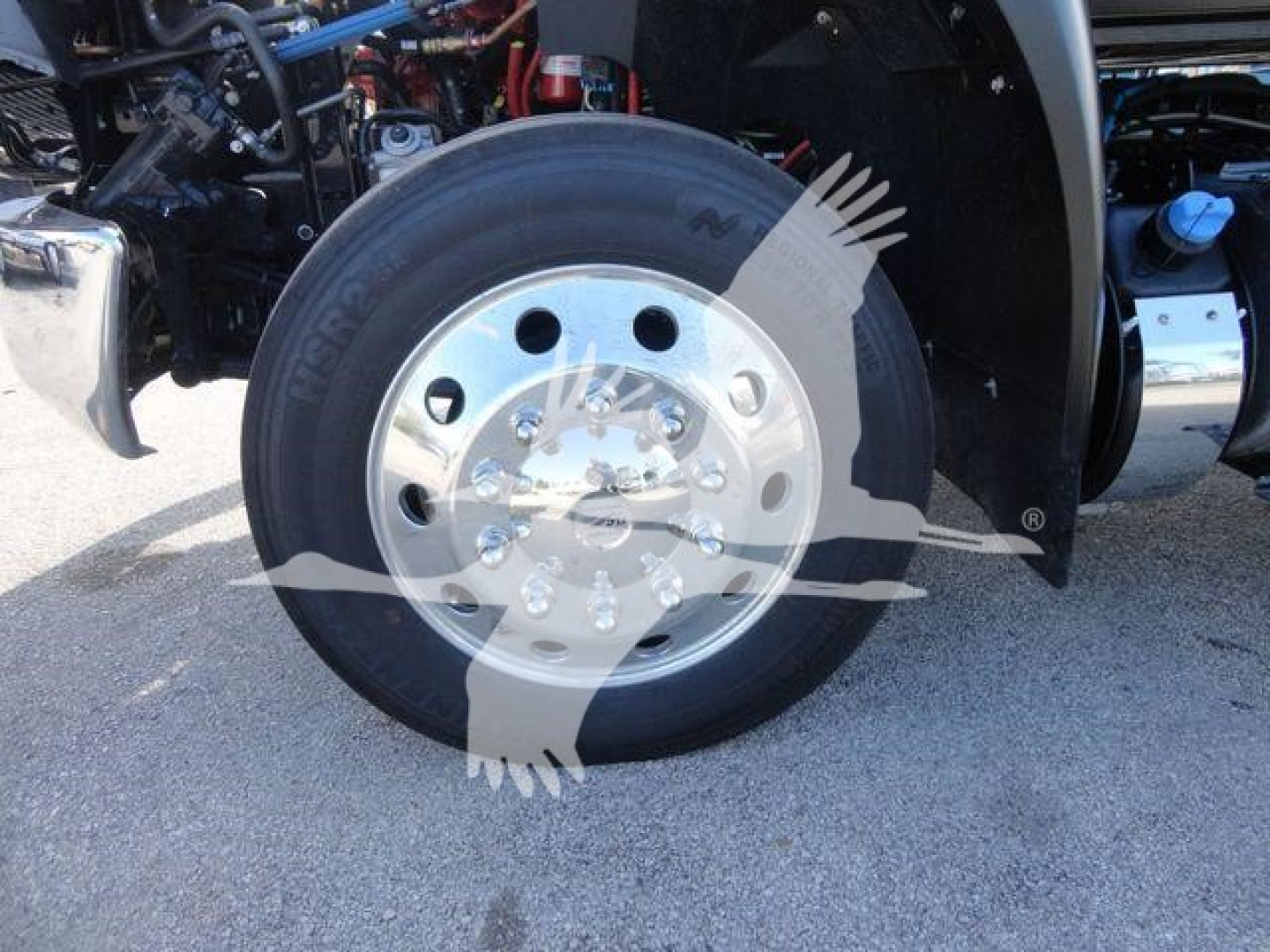 Truck front wheel and fender of a 2027 International MV rollback truck showing a polished aluminum wheel with multiple lug nuts and tire, nearby red engine components and blue cap visible; watermark graphic across image.