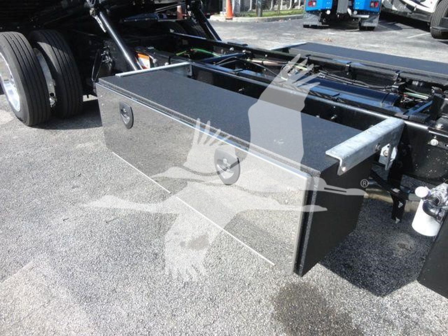 Truck chassis with a large rectangular metal toolbox mounted between the front and rear wheels, showing a metal latch on the toolbox door and a hydraulic/metal frame nearby.