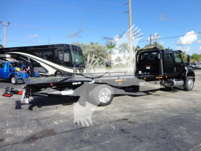 Flatbed tow truck on a lot, side view showing long wheelbase with black cab and extended flatbed; clear sky and other vehicles in background.