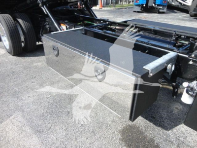 Truck chassis with a large rectangular metal toolbox mounted between the front and rear wheels, showing a metal latch on the toolbox door and a hydraulic/metal frame nearby.