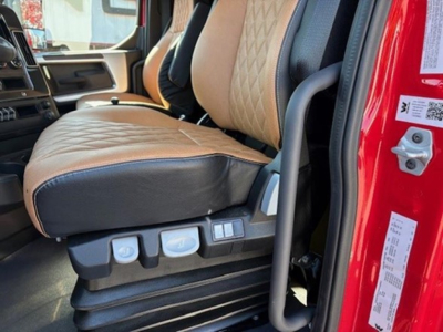 Truck cab interior showing tan leather seats with diamond stitching and controls on seat base. Door frame and red exterior visible.