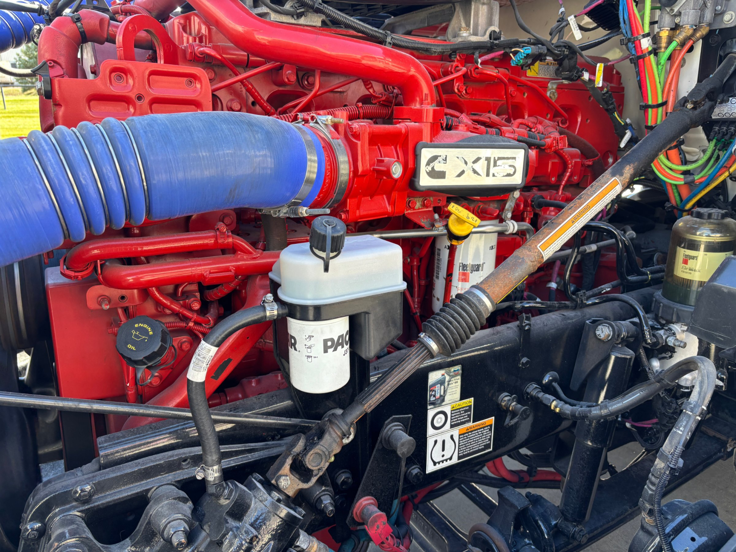 Kenworth T680 engine bay showing coolant reservoir, Dexron-style fluid lines, and red engine hardware with a white badge reading X15.