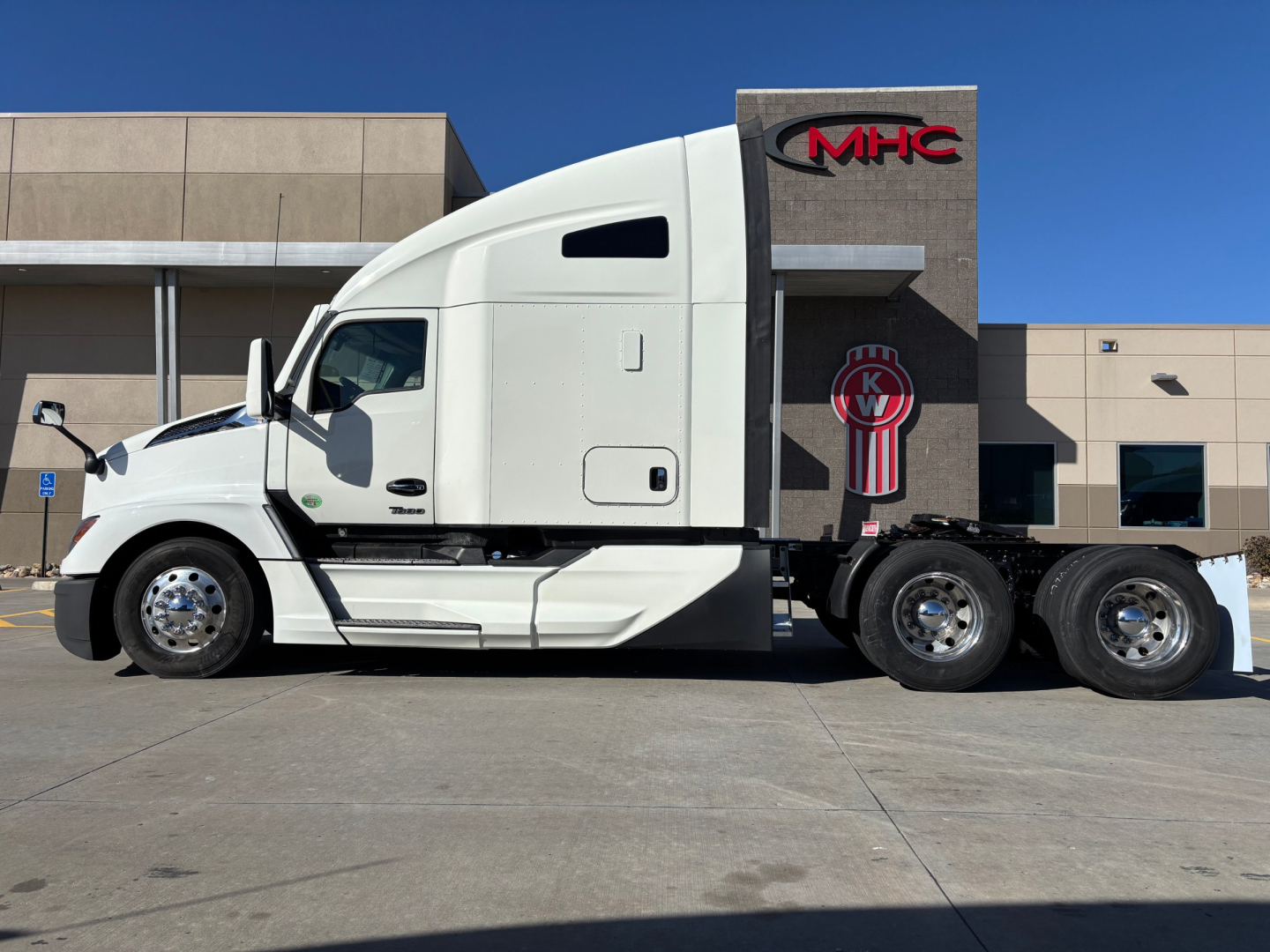 Kenworth T680 tractor in white, side profile view, sleeper cab, dual rear axles, badge 'MHC' on building behind.