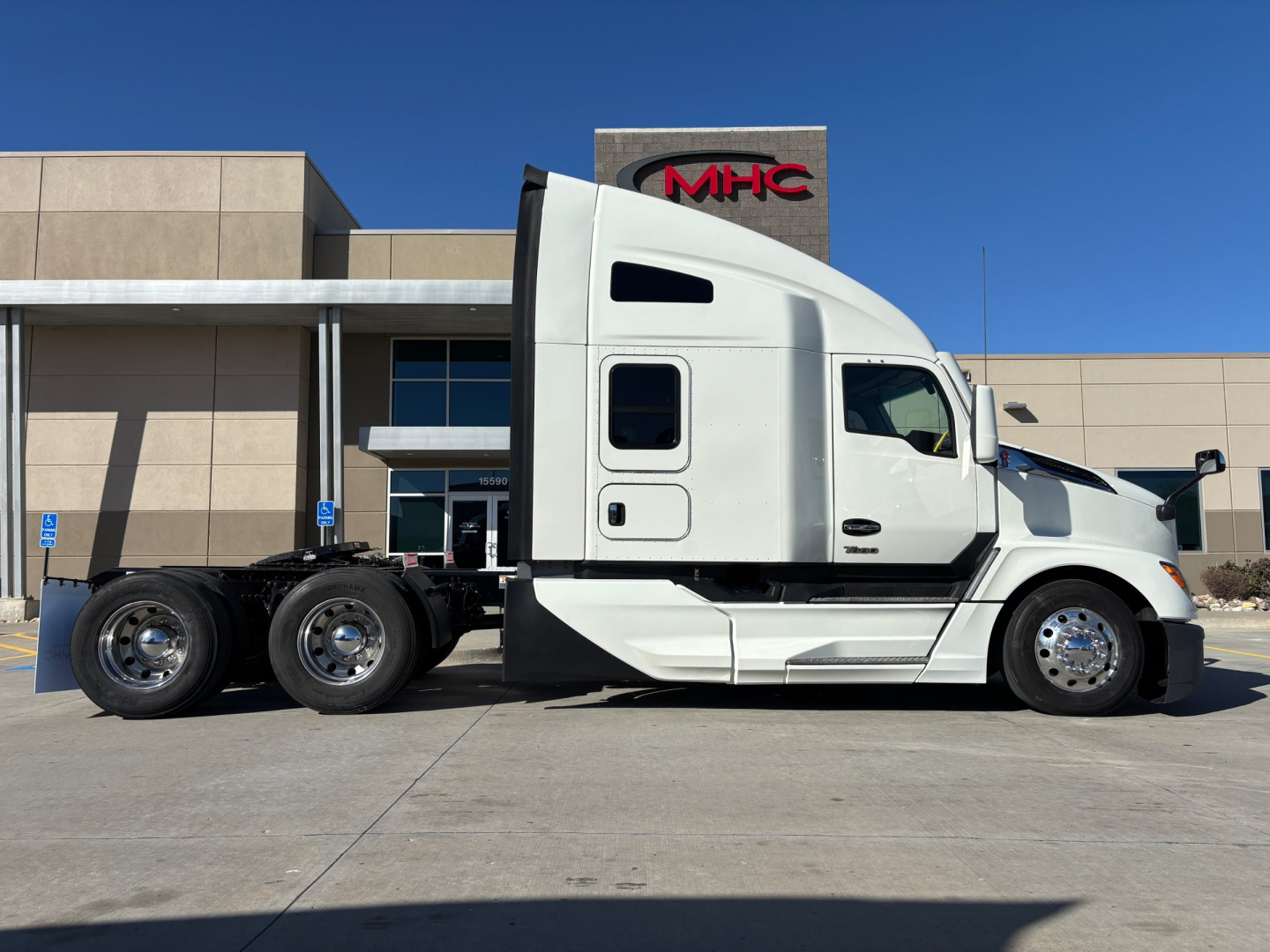 Kenworth T680 tractor side view showing sleeper cab, two rear axles, white exterior, and chrome wheels in front of dealership building.