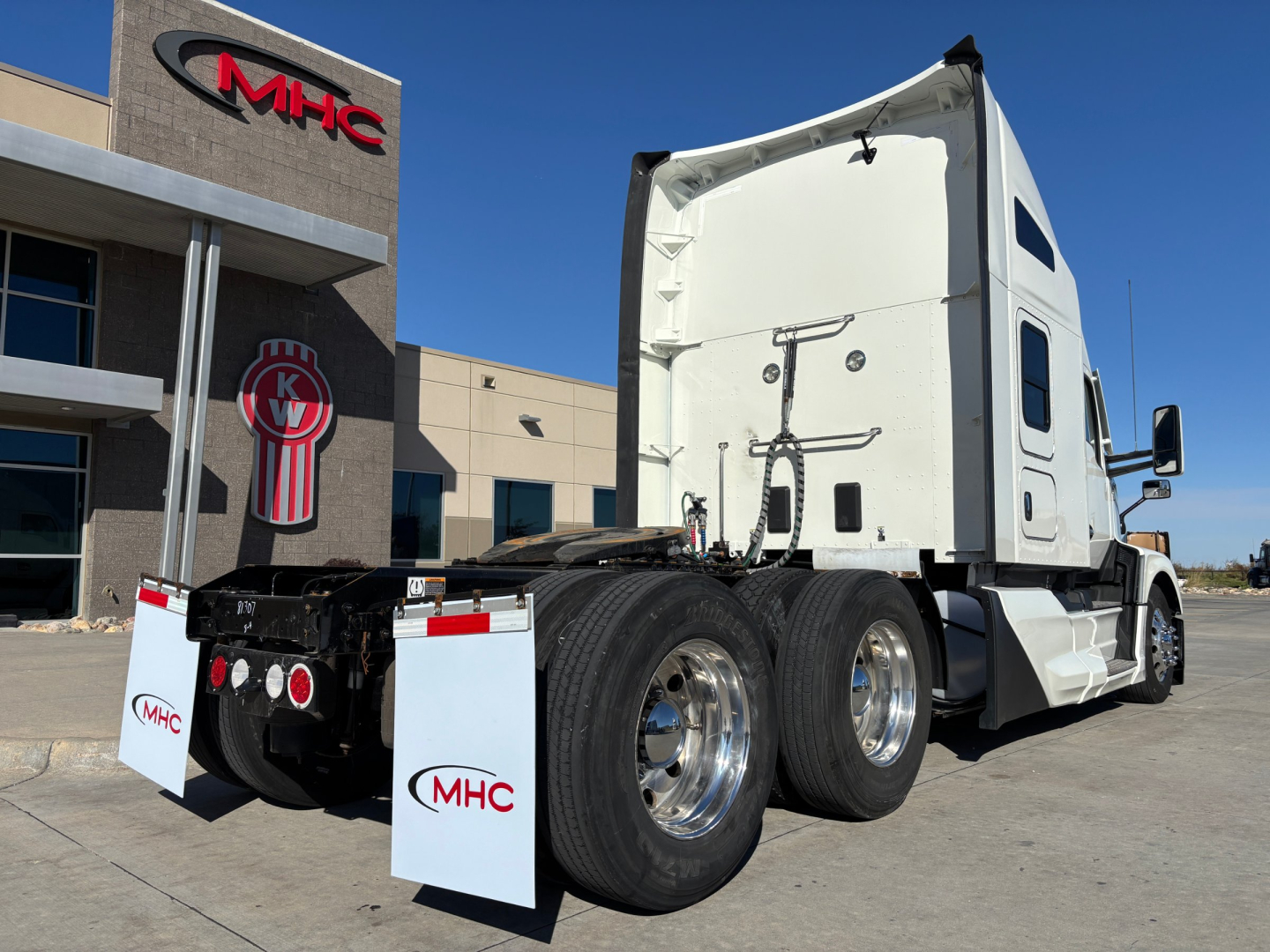 Kenworth T680 tractor rear-left angle showing triple rear axles, white cab, fifth wheel, and MHC dealership in background.