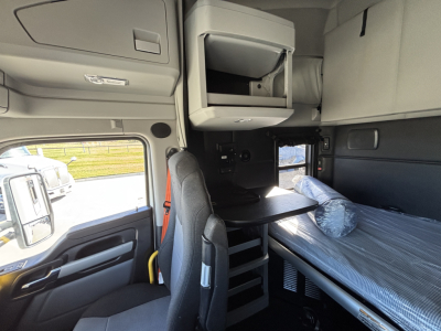 Cab interior of a Kenworth T680 sleeper truck showing bunk area with a plastic-wrapped mattress, overhead storage, and a small desk area; window and door visible.