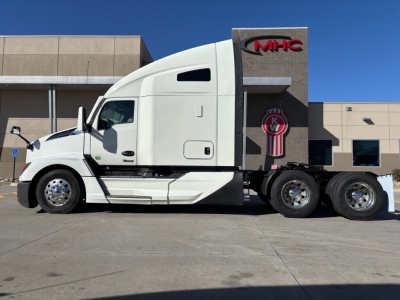 Kenworth T680 tractor in white, side profile view, sleeper cab, dual rear axles, badge 'MHC' on building behind.
