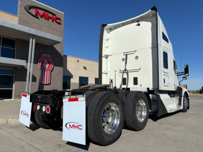 Kenworth T680 tractor rear-left angle showing triple rear axles, white cab, fifth wheel, and MHC dealership in background.