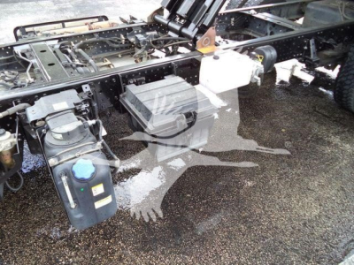 Isuzu NPR XD chassis showing coolant reservoir, hydraulic reservoir, and battery box with hoses and frame rail visible.