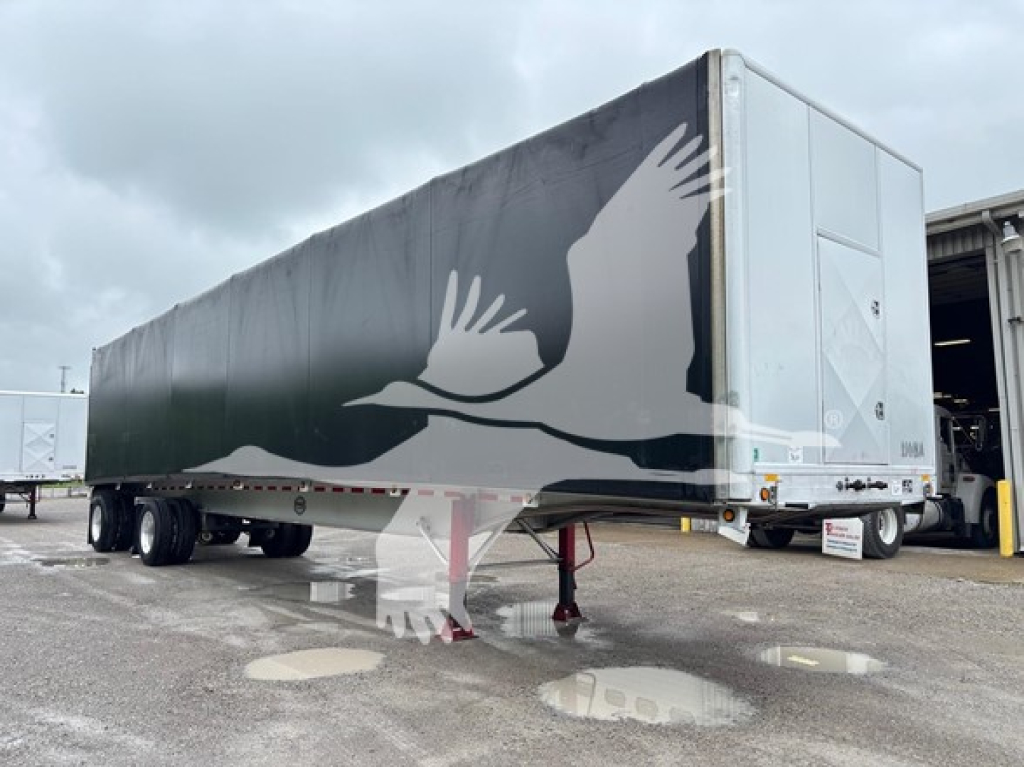 Trailer side view showing a large graphic of a flying crane on a dark exterior; the front of the trailer shows reflective panels and a white tail with support legs visible.