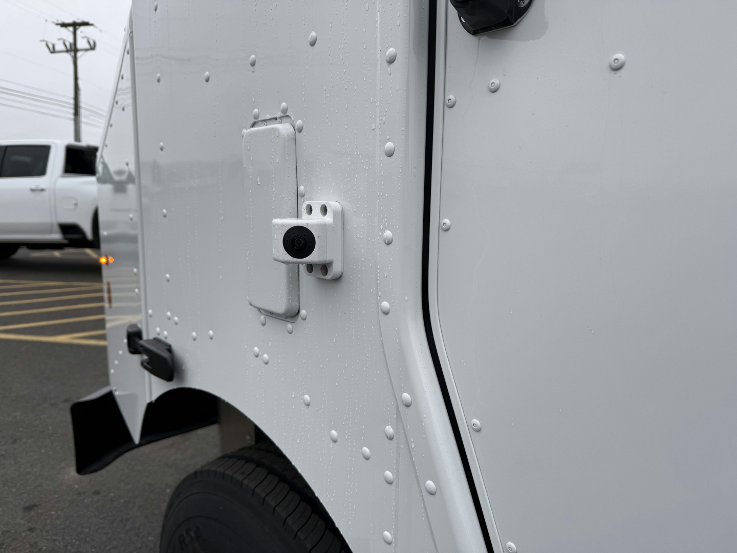 Harbinger S655 truck cab corner showing riveted metal panel, hinge, and a small black round camera or sensor on white body, treaded tire visible.