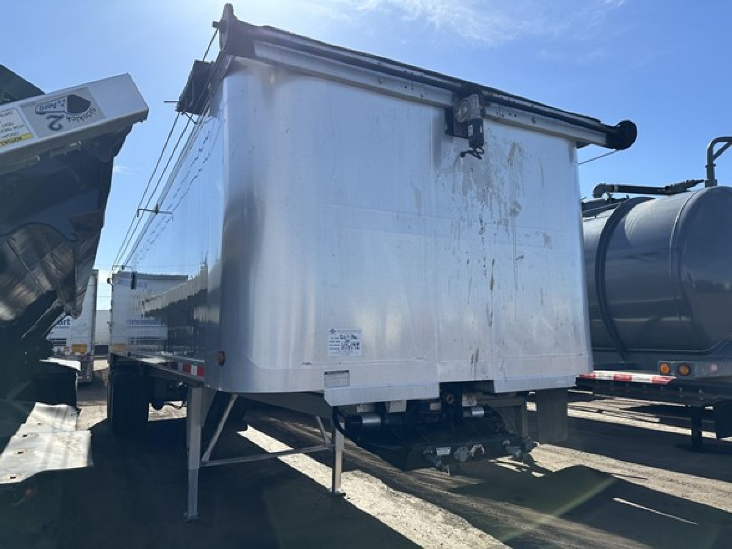 Dump trailer with aluminum box and dual rear legs, showing hitch and undercarriage; side panels with slight dents and a small label on the front corner.