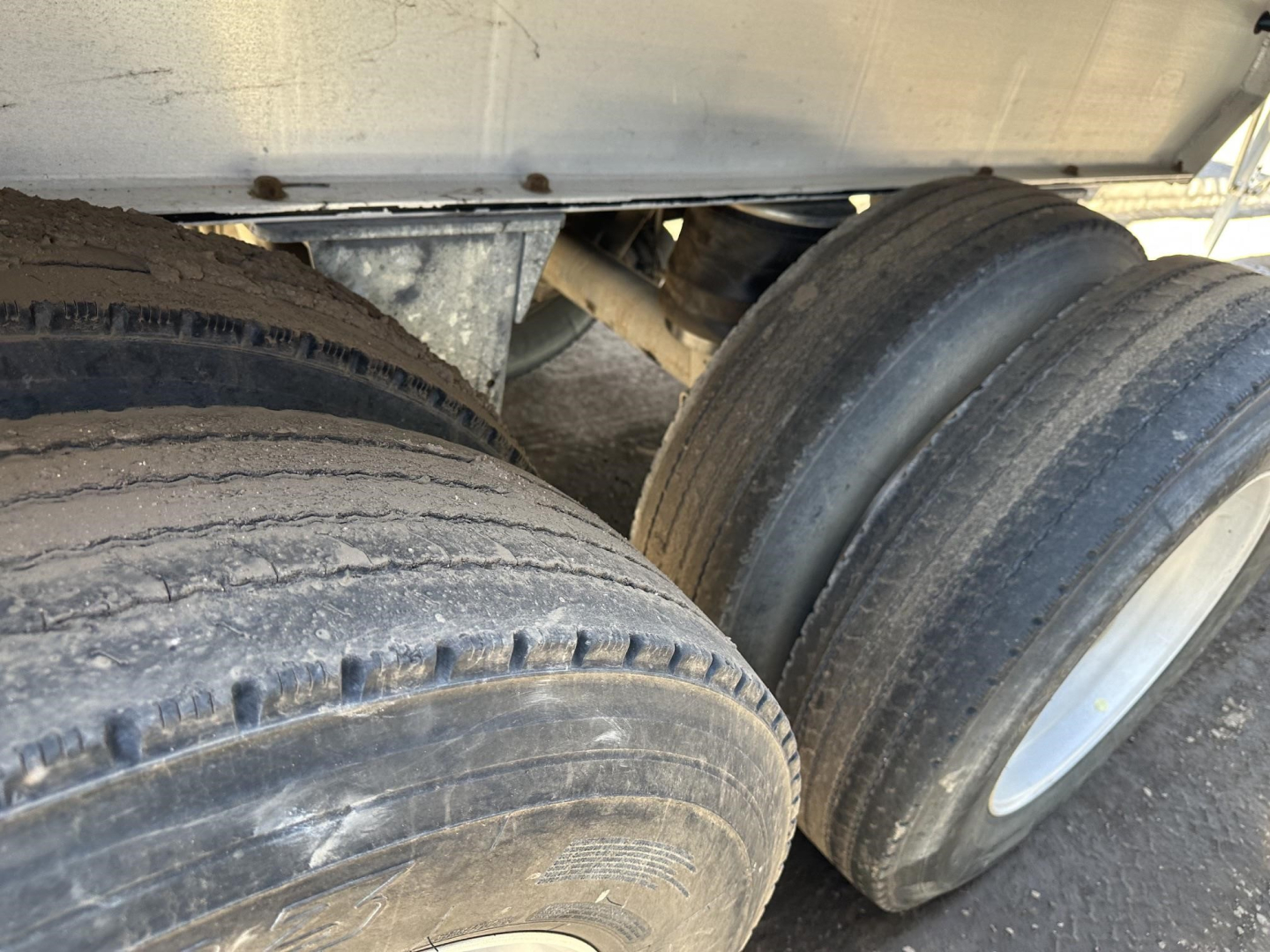 Dual trailer tires mounted under a dump trailer frame with mud and wear on tread.