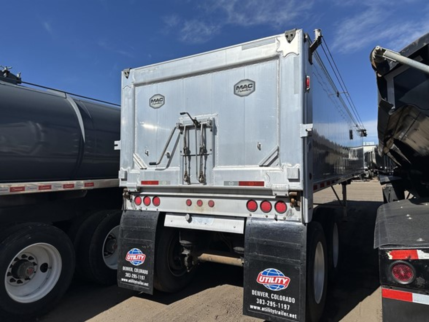 Rear view of a Mac dump trailer with dual axles, silver exterior, MAC logos on rear doors, tail lights, and mud flaps showing Utility and Colorado contact numbers.