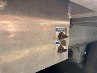 Dump trailer frame with two blue-labeled suspension control levers visible on a metal panel.