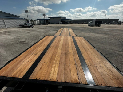 Trailer deck showing wooden drop deck with black steel frame and tandem axles in a dealership lot.